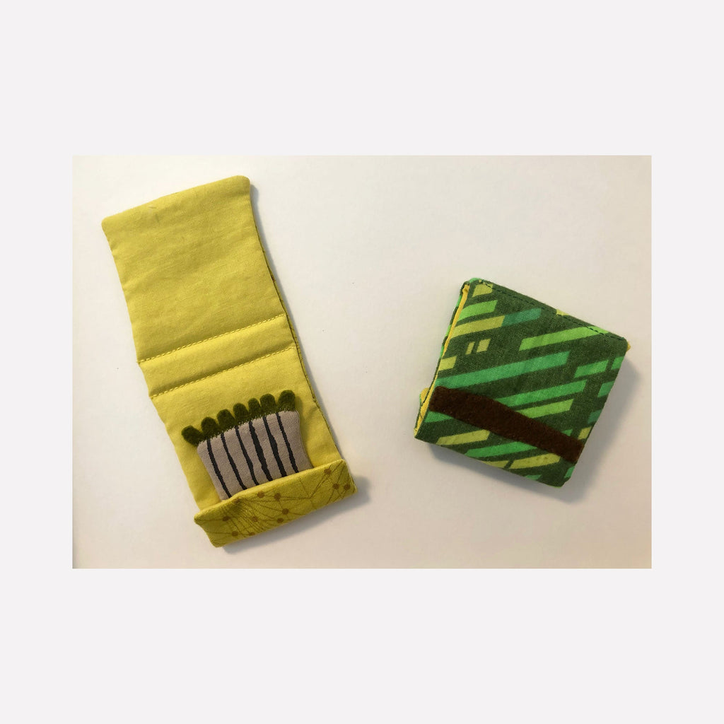 The artwork 'Matchbooks (Green)' by Ann Cofta features a hand-sewn fabric composition depicting two matchbooks - one open yellow matchbook displaying gray matches inside, and one closed matchbook with diagonal green striped pattern. Created with fabric, batting, and paint using appliqué technique, displayed in a shadowbox frame.