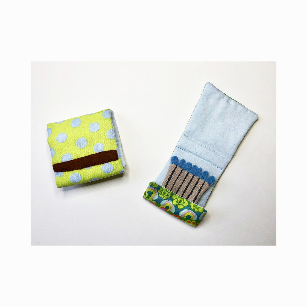 The artwork 'Matchbooks (Blue & Green)' by Ann Cofta features two fabric matchbook designs - one with yellow-green polka dots and brown stripe, another showing gray match sticks with blue tips against light blue fabric, adorned with a decorative green floral pattern trim. Hand-sewn fabric art with painted details. By Ann Cofta.