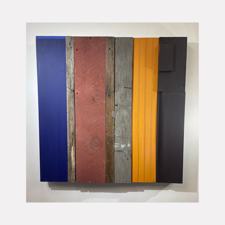 The artwork '36-01' by Simon Draper features vertical bands of painted wood in contrasting colors: deep navy blue, weathered rose, distressed gray, vibrant yellow, and matte black. The assemblage combines smooth and textured surfaces, creating a geometric abstract composition with rustic elements, measuring 36x36 inches.