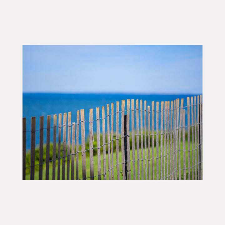 The artwork 'A Trip to Montauk' by Garrett Carroll captures a serene coastal scene featuring a weathered wooden beach fence against a vibrant blue ocean backdrop. The composition showcases soft focus photography highlighting the natural textures of the slatted fence, green coastal grass, and the endless horizon where azure waters meet clear skies.