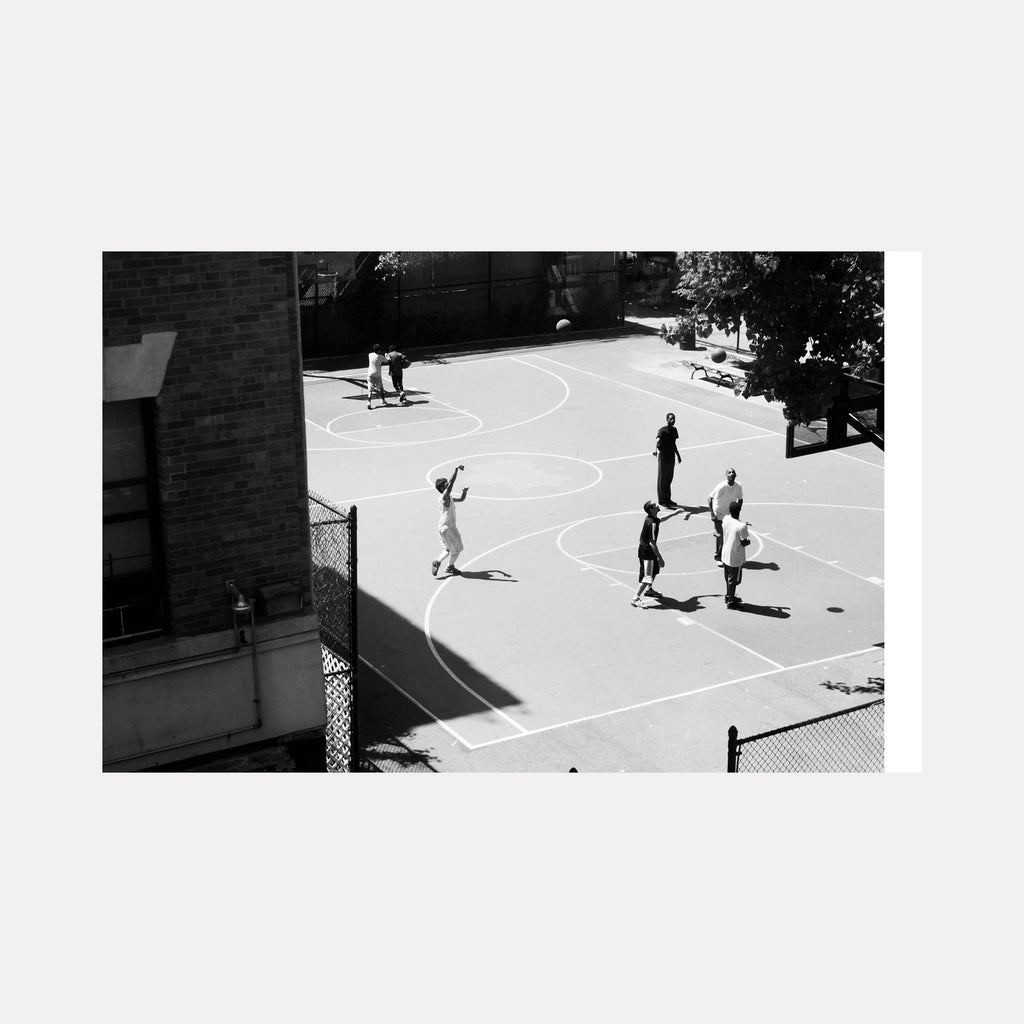 The artwork 'Ball is Life' by Cavier Coleman captures a black and white photograph of an outdoor basketball court scene. The composition shows players on a sunlit concrete court with crisp white painted lines forming multiple circles. The geometric patterns of the court markings contrast sharply against the dark brick building and chain-link fence borders, creating a striking urban sports landscape.