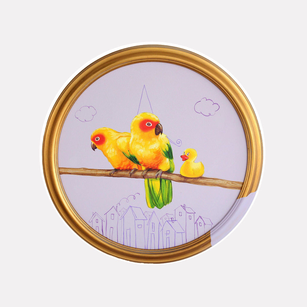 The artwork 'Birthday Surprise' by Carlos Gamez de Francisco features two vibrant yellow sun conures perched on a brown branch, accompanied by a small rubber duck. The birds are rendered in bright acrylics against a soft lavender background, with sketched clouds and a whimsical cityscape silhouette below. The circular composition is framed in an elegant gold round frame. 16x16 inches acrylic on canvas.