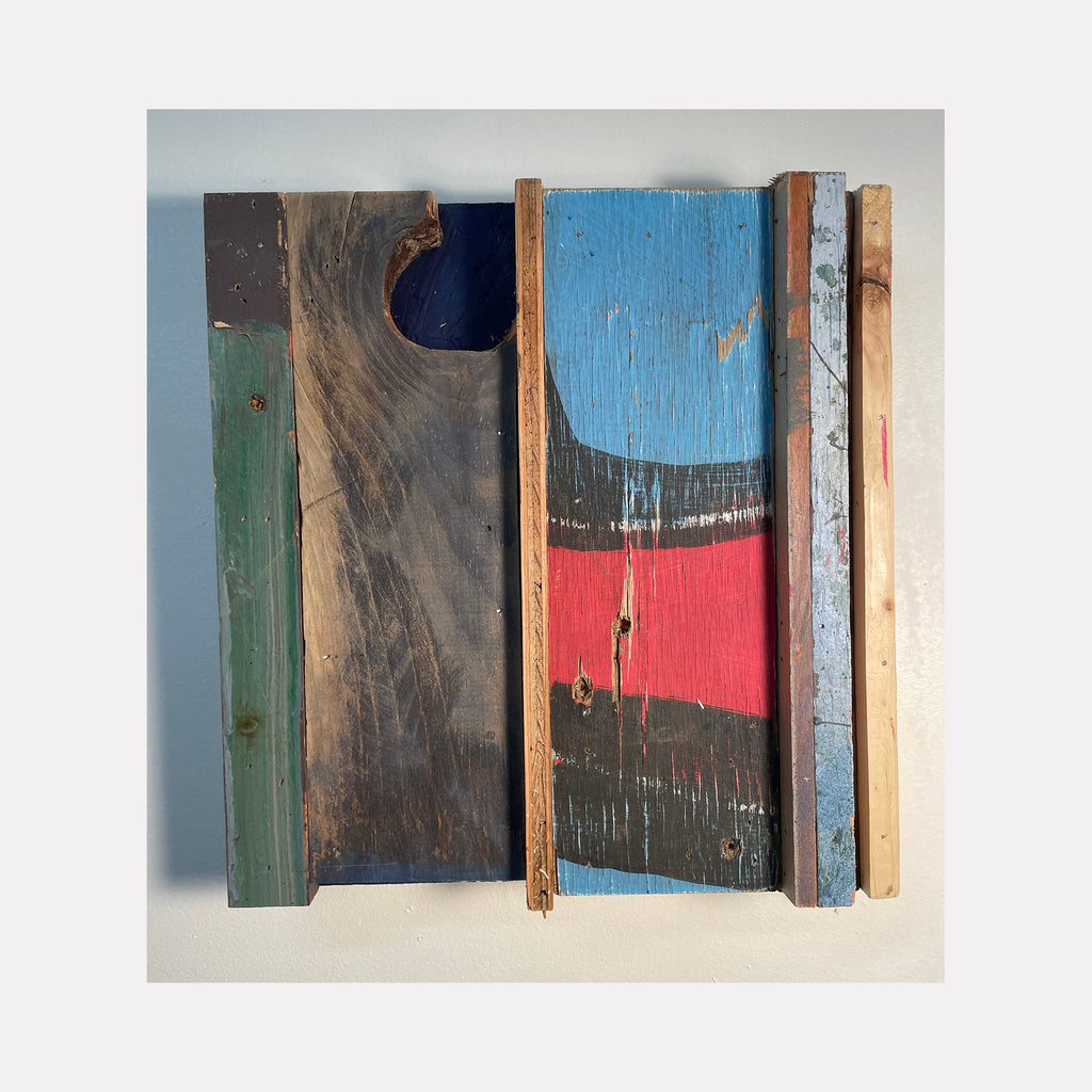 The artwork 'Brecon' by Simon Draper features a rustic wooden assemblage combining weathered planks in contrasting colors. Vertical panels showcase distressed blue, coral red, dark brown, and sage green painted surfaces, creating an abstract geometric composition. The textured wood grain and worn paint give the 12x12 inch piece a vintage, coastal-inspired character.