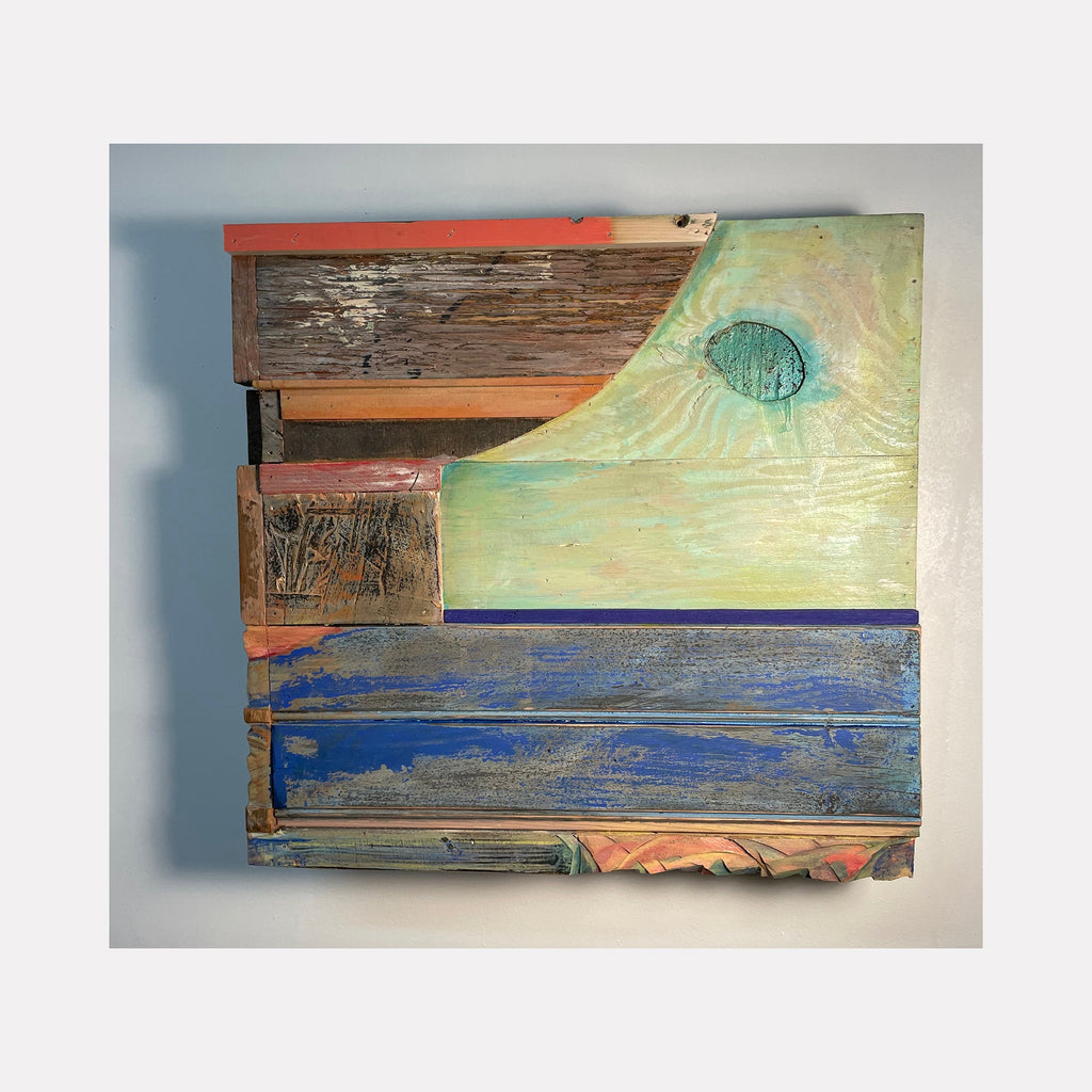 The artwork 'Build Where You Go' by Simon Draper features a geometric abstract composition of weathered wood pieces assembled in layers. The piece combines mint green, coral, and deep blue painted sections with distressed natural wood textures. A circular turquoise element appears in the upper right against a pale green surface, creating a sun-like focal point.