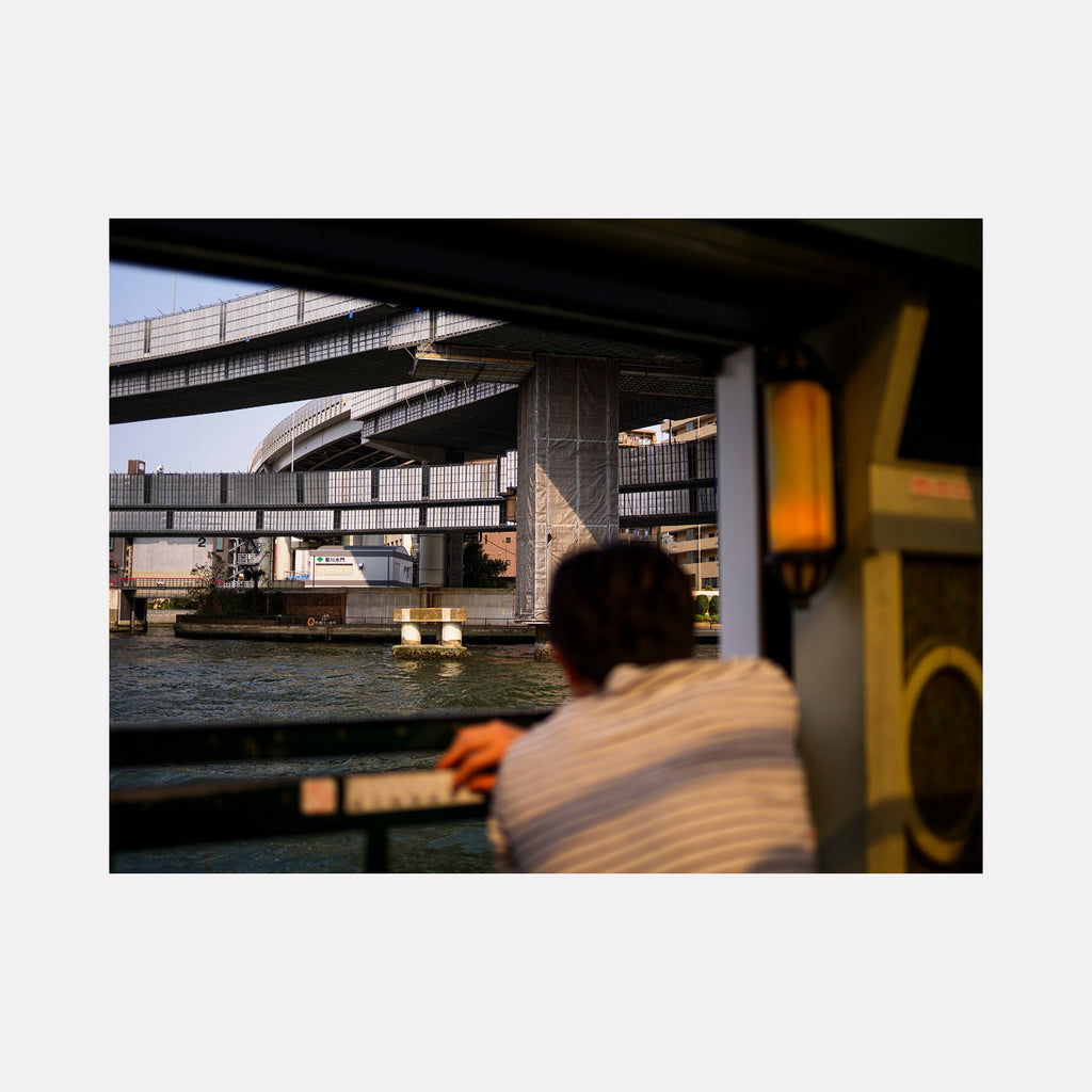 The artwork 'Calm Traveler' by Garrett Carroll captures a serene urban waterfront scene, featuring multi-level concrete highway overpasses crossing a blue waterway. A warm amber lantern illuminates the foreground, while industrial architecture creates striking geometric patterns against an overcast sky. The photograph's composition emphasizes the contrast between hard urban infrastructure and tranquil water reflections.