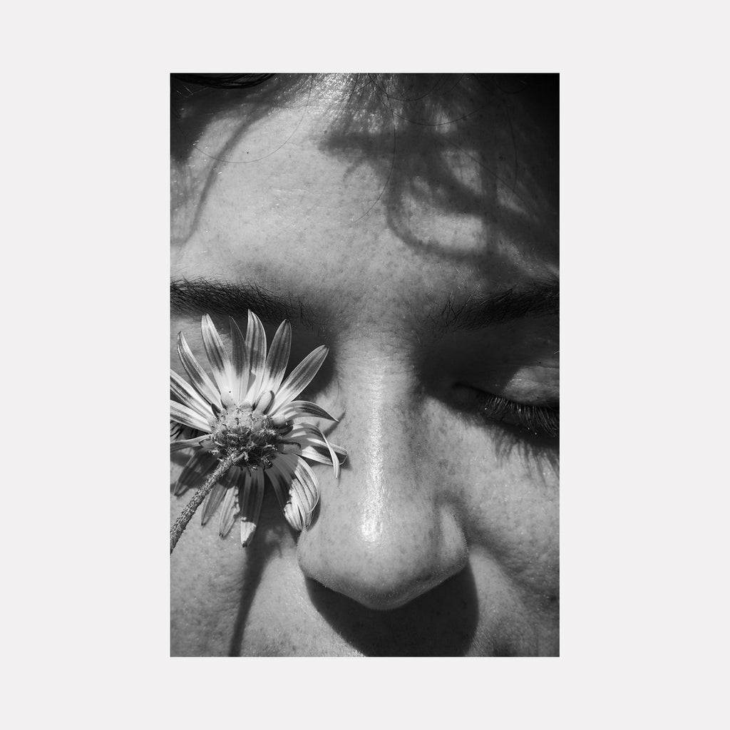 The artwork 'Communion III' by Eunice Pais features a dramatic black and white photograph capturing an intimate composition of a delicate daisy flower against skin. The monochromatic treatment emphasizes light and shadow, with the flower's detailed petals creating a striking contrast. A macro-style artistic photography that explores texture and natural forms. By Eunice Pais.