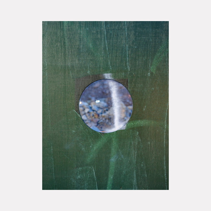 The artwork 'Construction Fence Series-6' by Garrett Carroll captures a circular peephole view through a weathered green construction fence. A blurred, abstract scene in cool blue tones is visible through the circular opening, creating an intriguing contrast against the textured, forest-green surface of the fence. The photograph's composition plays with perspective and depth through architectural elements.