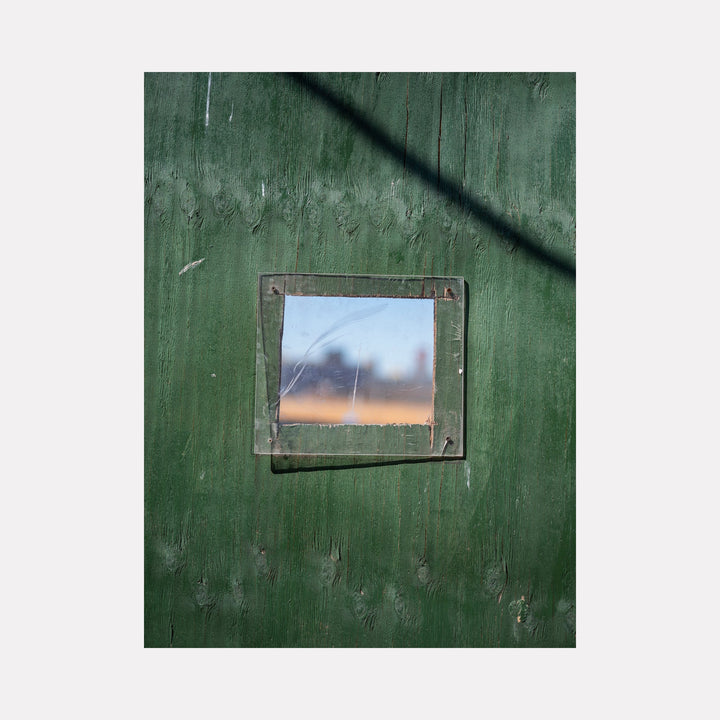 Construction Fence Series-8 by Garrett Carroll captures a unique perspective through a small square window in a weathered green construction fence. The photograph features a metallic-framed opening revealing a hazy sunset sky, creating a striking contrast between industrial materials and natural light. The textured, forest-green fence surface dominates the composition.