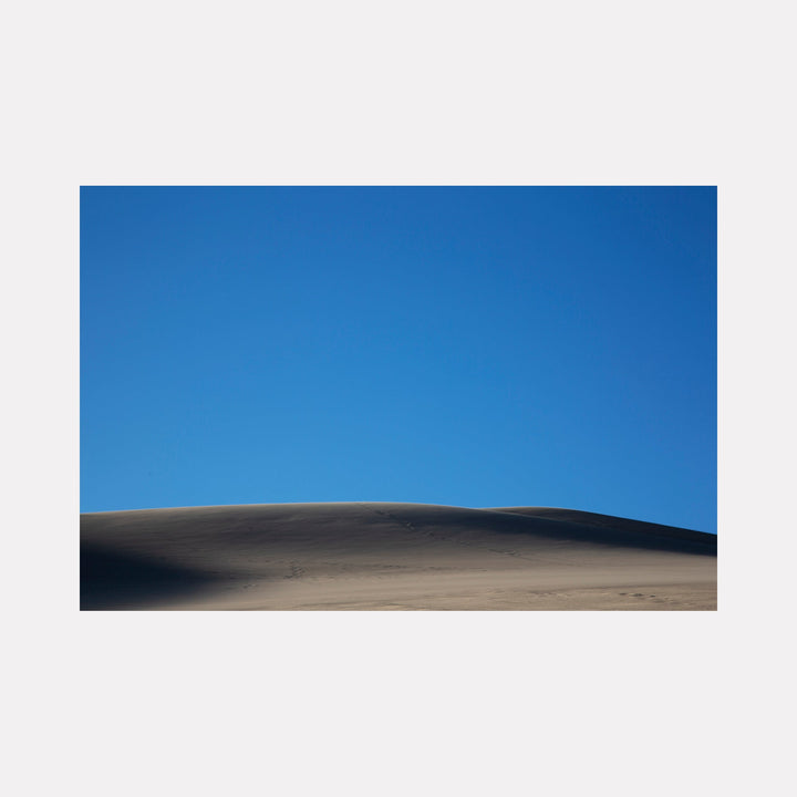 The artwork 'Dune' by Cavier Coleman captures a minimalist desert landscape photograph featuring smooth, rolling sand dunes against a pristine azure blue sky. The composition emphasizes the natural curves and shadows of the desert terrain, creating a striking contrast between the warm sandy tones and the deep blue horizon. Fine-art photography print, 24x36 inches.