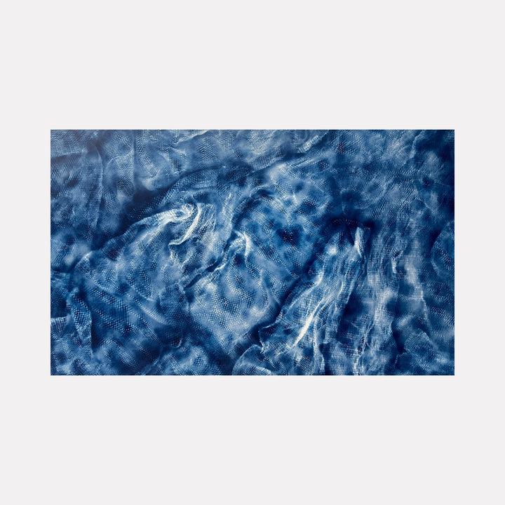 The artwork 'Entangled' features swirling patterns of deep ocean blues and white streaks creating a dynamic, fluid composition reminiscent of turbulent waters or cloudy skies. The spray paint technique produces ethereal textures and movement across the large-scale canvas, with varying intensities of indigo and cerulean hues merging seamlessly. By Gina Iannucci.