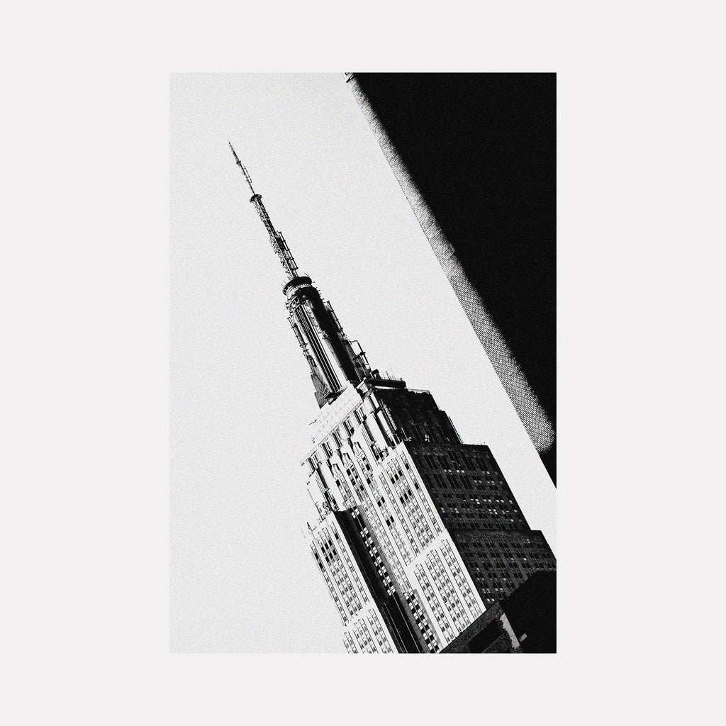 Empire State of Mind by Cavier Coleman: Dramatic black and white photographic capture of the Empire State Building's iconic spire and upper facade, shot from a striking angular perspective. The architectural masterpiece emerges from stark contrast between light and shadow, showcasing Art Deco details and the building's majestic presence against a bright sky.