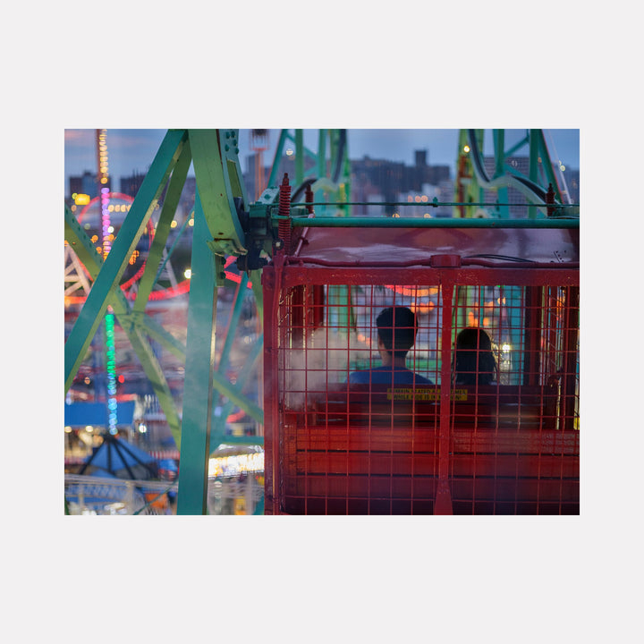 The artwork 'Ferris Wheel Ride at Dusk' by Garrett Carroll captures a vibrant carnival scene, featuring a red passenger cabin against green metal support beams. Colorful lights sparkle in the twilight background, creating a magical atmosphere. The composition showcases carnival's evening ambiance with glowing blue, pink, and yellow illumination reflected across the frame.