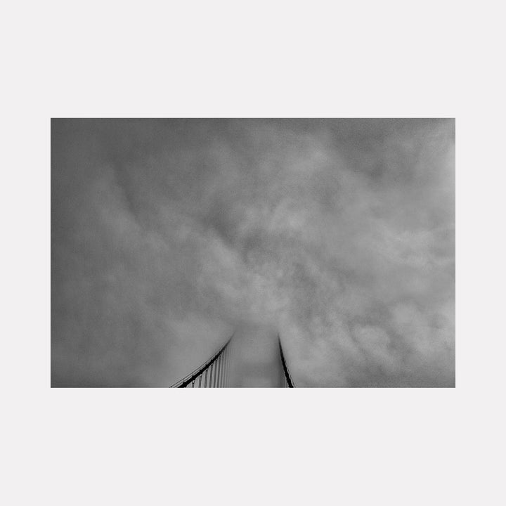 The artwork 'Golden Gate Bridge' by Neil Shapiro features a dramatic black and white perspective of bridge cables rising into foggy skies. The minimalist composition captures the iconic suspension bridge's vertical lines disappearing into moody cloud cover, printed on textured Hahnemühle paper with rich monochromatic tones.