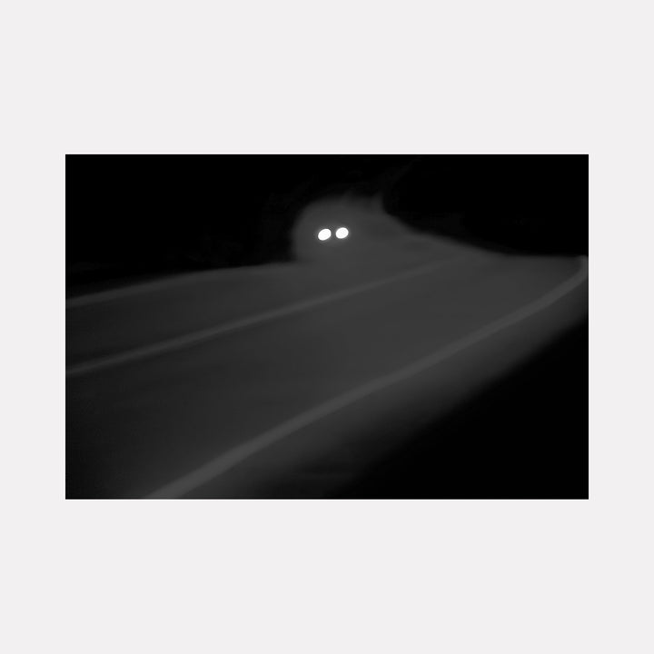 The artwork 'High Beams' by Neil Shapiro captures a minimalist black and white photograph of car headlights emerging from darkness on a curved road. The composition features two bright circular lights against a misty or foggy background, creating a dramatic contrast between light and shadow. Printed on textured Hahnemuhle paper, the image evokes a sense of mystery and nocturnal atmosphere.