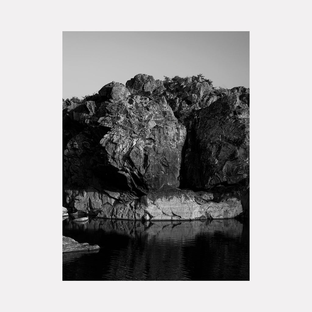 The artwork 'Hike to the Falls' by Garrett Carroll captures a dramatic black and white landscape photograph featuring rugged cliff faces reflected in still waters. Jagged rock formations dominate the composition, with deep shadows and textures emphasizing the natural geometric patterns. The tranquil water surface creates a mirror-like effect, doubling the visual impact of the rocky terrain.