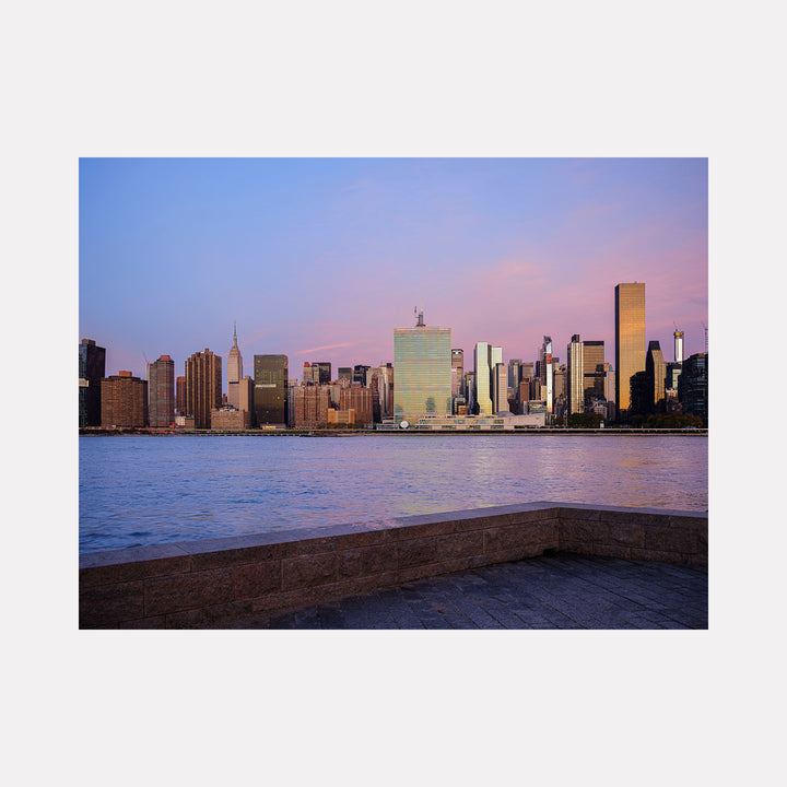 The artwork 'Hunters Point Sunrise' by Garrett Carroll captures Manhattan's iconic skyline at dawn, featuring gleaming skyscrapers reflected in East River's calm waters. Pink and purple hues paint the morning sky, while a concrete embankment frames the foreground, creating a serene urban landscape photographed from Queens.