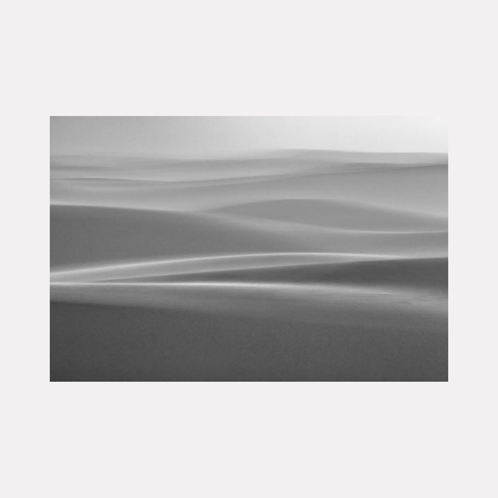 The artwork 'Magical Dunes' by Neil Shapiro captures ethereal desert landscape in monochromatic tones. Smooth, undulating sand dunes create flowing lines across the composition, with subtle gradients of light and shadow enhancing the dreamy atmosphere. The minimalist black and white photography printed on textured Hahnemuhle paper emphasizes the abstract, sculptural quality of the natural formations.