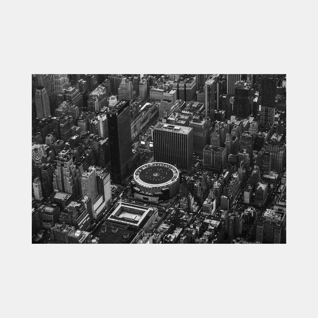 The artwork 'MSG' by Cavier Coleman captures a dramatic aerial black and white photograph of Madison Square Garden and surrounding Manhattan skyscrapers. The iconic circular arena stands prominently amid the urban landscape of towering buildings, creating a striking architectural contrast in the heart of New York City's skyline. 24x36 inches photographic print.