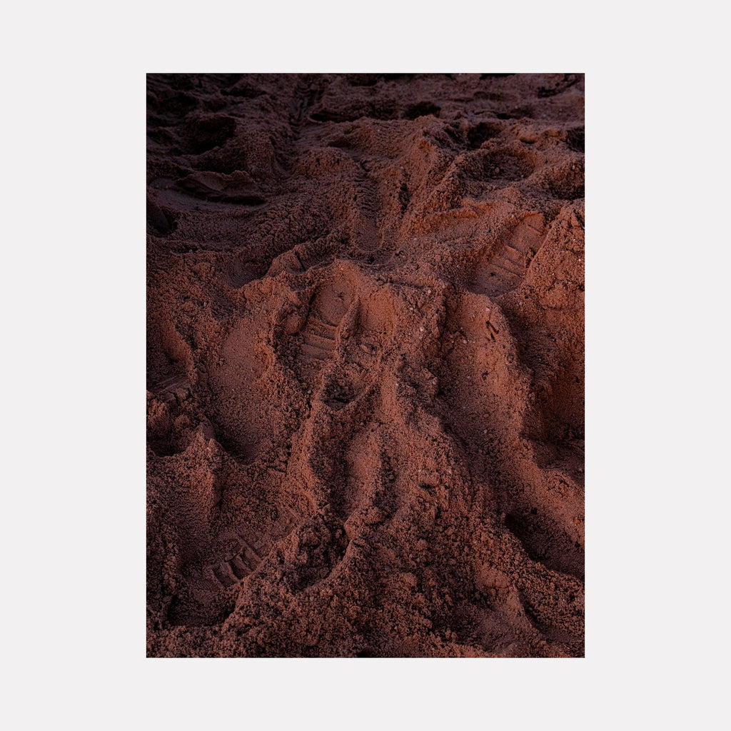 The artwork 'Morning Beach' by Garrett Carroll captures intricate textures of deep reddish-brown sand with natural ripples and footprints creating an abstract pattern. The close-up photography showcases the rich, earthy tones and granular texture of beach sand illuminated in morning light, displaying both depth and dimensional qualities. Photography by Garrett Carroll.