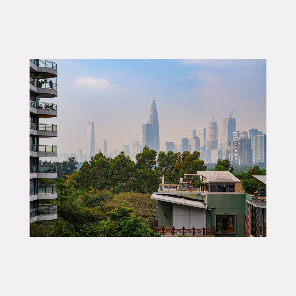 The artwork 'Overlooking at Your Home' by Garrett Carroll captures a modern urban landscape with towering skyscrapers rising above lush green trees. The photograph features contemporary high-rise buildings with balconies in the foreground, while distant glass and steel skyscrapers pierce a soft blue sky. A verdant tree line creates a natural barrier between residential areas and the city skyline.