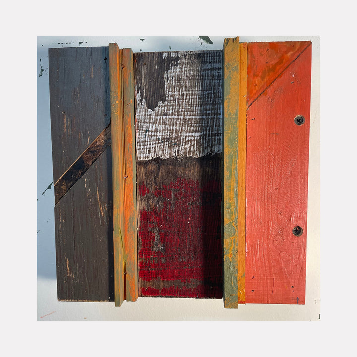 The artwork 'Phoenix' by Simon Draper features a geometric wood assemblage with three distinct vertical panels in charcoal gray, deep crimson, and vibrant orange. Textural elements include weathered wood grain, scraped paint layers, and exposed raw surfaces, creating a rustic abstract composition measuring 12x12 inches.