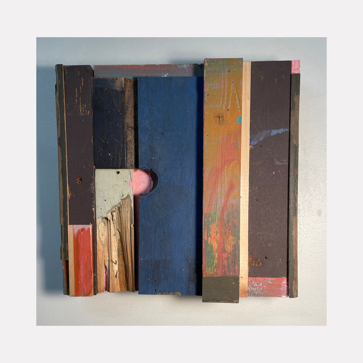 The artwork 'Prebuilt' by Simon Draper features a geometric assemblage of weathered wooden planks arranged vertically in varying widths. The composition combines deep navy blue, rustic brown, coral pink, and natural wood tones, creating a textural abstract pattern. The distressed painted surfaces and raw wood edges add industrial character to this 12x12 inch wall-mounted piece.