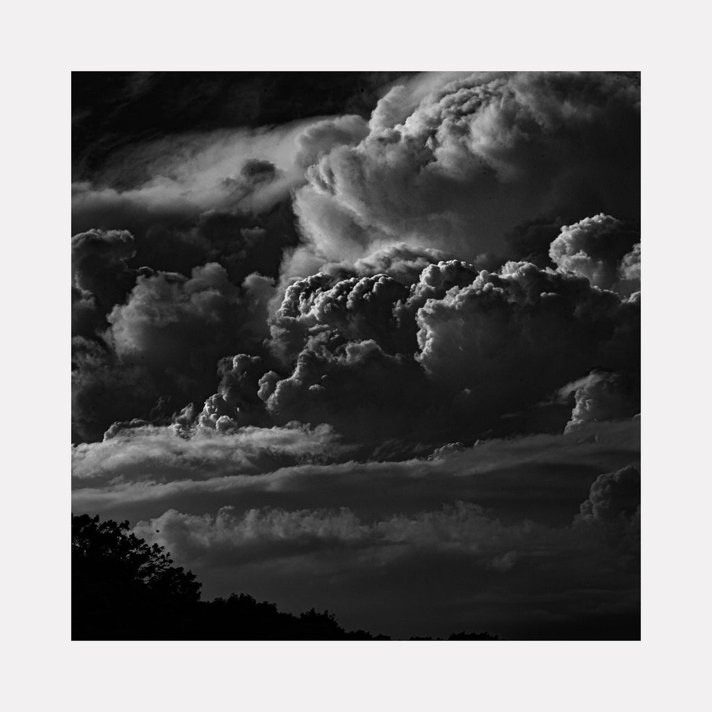 The artwork 'Running Clouds' by Cavier Coleman captures dramatic storm clouds in black and white photography. Billowing cumulus formations dominate the composition, creating striking contrast against a dark sky. Silhouetted trees line the bottom edge, while textured cloud layers build upward with powerful dimensional depth. Fine art photographic print measuring 24x36 inches.