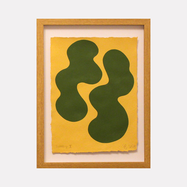 The artwork 'Synergy II' by Shadia Sabagh features two organic, flowing green shapes set against a vibrant yellow background, framed in light wood. The abstract composition showcases fluid, biomorphic forms reminiscent of natural elements, painted in acrylic on handmade cotton paper with deckled edges, creating a harmonious balance between color and form.