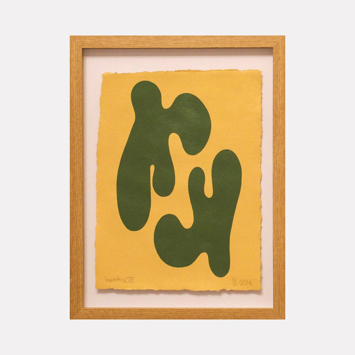 The artwork 'Synergy III' by Shadia Sabagh features abstract organic shapes in deep forest green against a vibrant yellow background. The minimalist composition displays flowing, biomorphic forms reminiscent of natural elements, painted in acrylic on handmade cotton paper, framed in light oak wood.