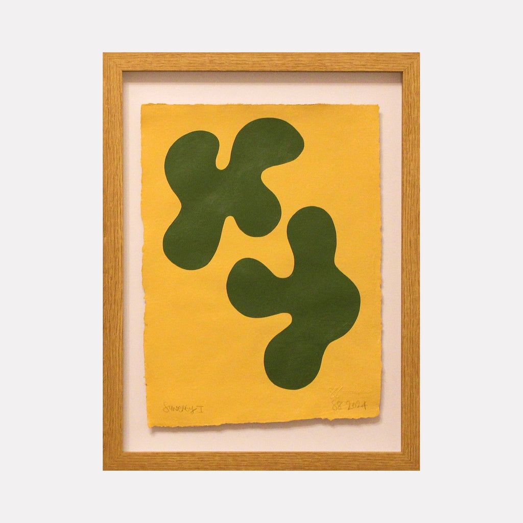 The artwork 'Synergy I' by Shadia Sabagh features two organic, amoeba-like dark green shapes floating on a vibrant yellow background. The abstract composition is rendered in acrylic paint on deckle-edge cotton paper, framed in light wood. The minimalist design creates a dynamic interplay between positive and negative space.