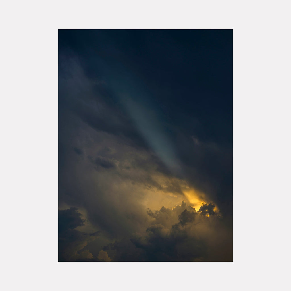 The artwork 'Saying Bye to Friends' by Garrett Carroll captures a dramatic skyscape where golden sunlight pierces through dark storm clouds, creating a ethereal contrast between deep navy blues and warm amber hues. The photograph showcases billowing cumulus clouds illuminated from behind, with streaking rays of light breaking through the moody atmospheric composition.