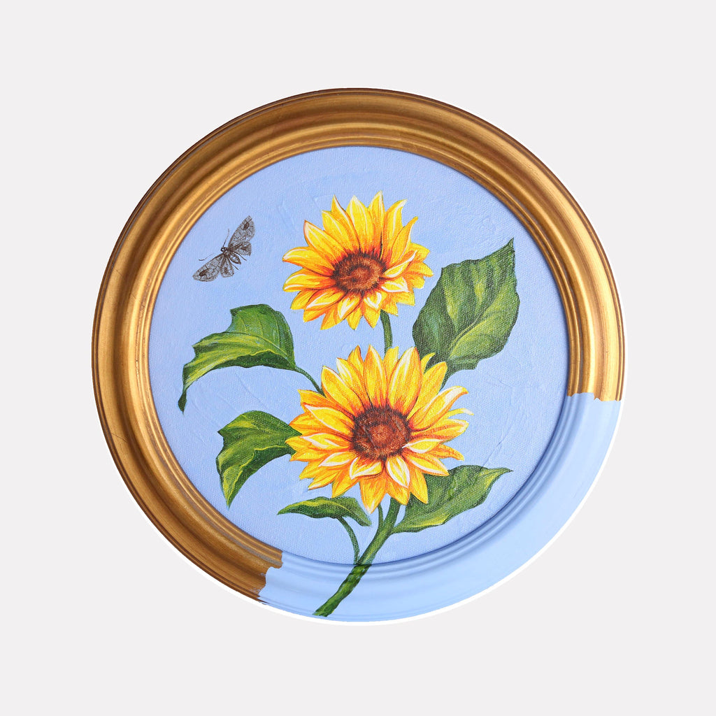 The artwork 'Sunflowers' by Carlos Gamez de Francisco features vibrant yellow sunflowers with rich brown centers against a serene light blue background, framed in a circular golden frame. Two blooming sunflowers with lush green leaves are accompanied by a delicate moth, painted in acrylic on a 10x10 inch canvas with realistic botanical detail.