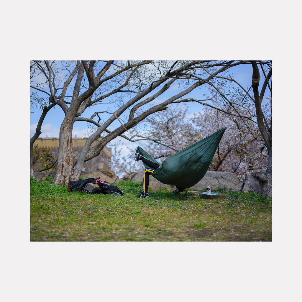 The artwork 'Suspended in Spring' by Garrett Carroll captures a serene outdoor scene featuring a dark green hammock suspended between trees on a grassy hillside. Bare branches frame the composition against a soft blue sky, while spring blossoms add delicate touches of color. A backpack rests nearby on the ground, creating a peaceful wilderness camping atmosphere in this fine art photography piece.