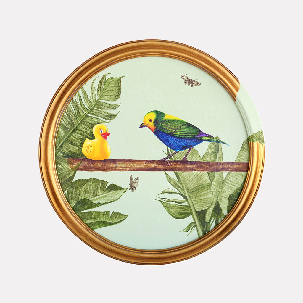 The artwork 'The Conversation' by Carlos Gamez de Francisco features a whimsical scene of a yellow rubber duck facing a vibrant blue and yellow tropical bird perched on a brown branch, surrounded by lush green banana leaves. Small moths flutter in the mint green background, all set within a circular gold frame. Acrylic on canvas, 16x16 inches.