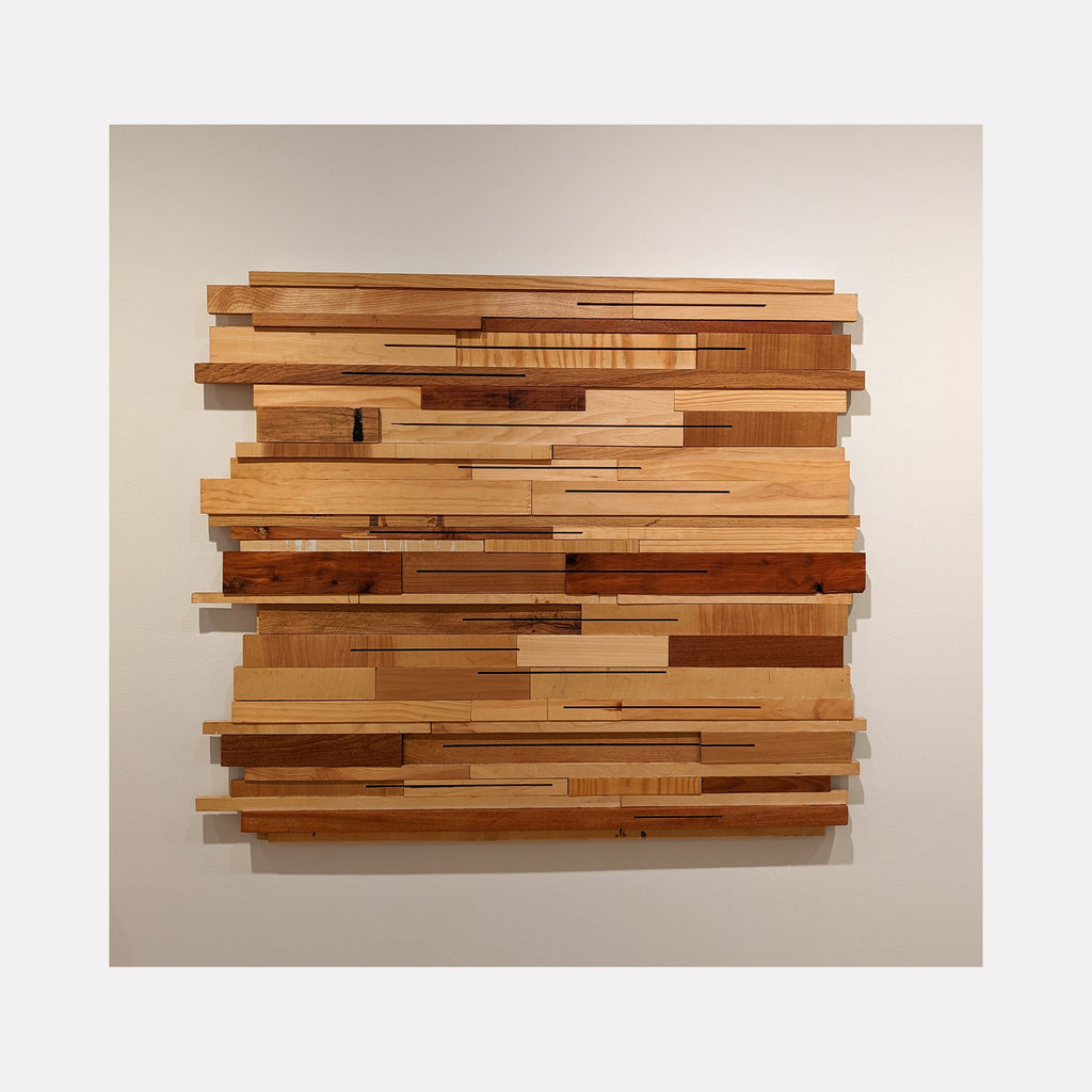 The artwork 'These Woods Were Trees I' by Ryan Frank features a geometric wall sculpture composed of layered wooden strips in varying lengths and tones. Multiple wood species including birch, mahogany, and cedar create a dynamic horizontal pattern with natural grain textures and warm honey to deep brown hues. The pieces are artfully arranged to suggest a deconstructed forest landscape.