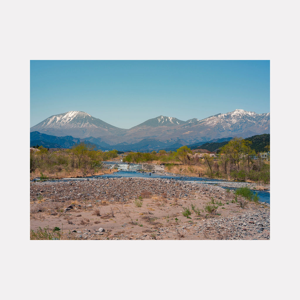 Tochigi Prefecture Mountainside by Garrett Carroll: A serene landscape photograph capturing snow-capped mountain peaks against a clear blue sky, with a winding river cutting through rocky terrain. Spring vegetation dots the riverbanks, while distant mountains showcase dramatic elevation changes and rugged topography in the Japanese countryside.