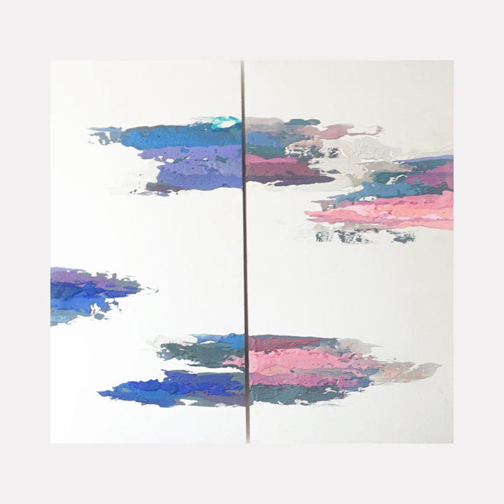 The artwork 'Twin Flames' by Sofia Echa features ethereal abstract brushstrokes in blues, pinks, and purples floating across a pristine white background, divided by a central metallic line on stainless steel. The watercolor-like acrylic strokes create a dreamy, cloud-like formation, suggesting movement and fluidity across the diptych composition.