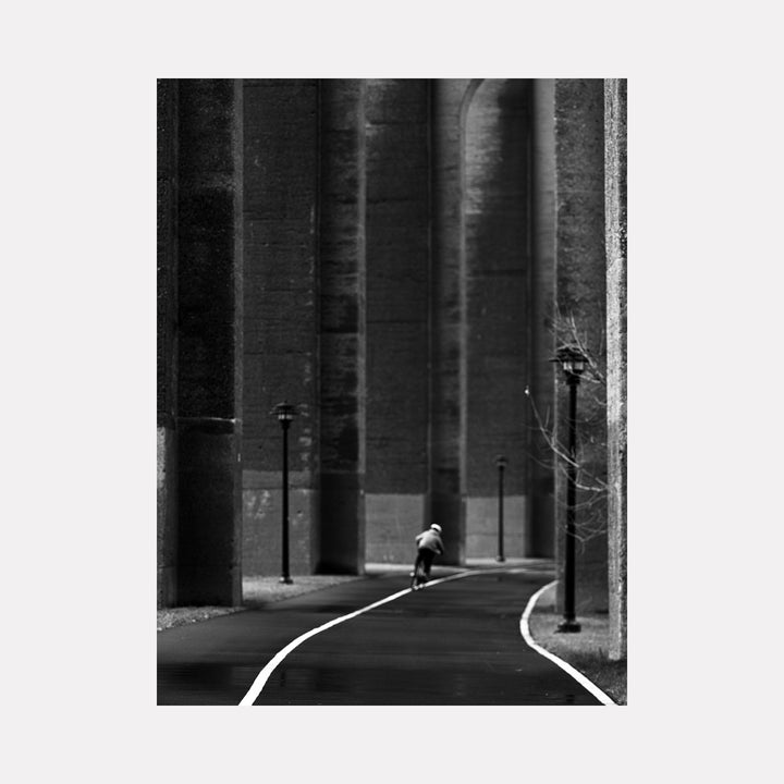 The artwork 'Underneath Hells Gate - Series-2' by Garrett Carroll captures a dramatic black and white photograph of a solitary figure on a curved pathway beneath towering concrete bridge pillars. Stark contrast emphasizes the monumental scale, with street lamps creating a rhythmic pattern along the winding path. The composition conveys urban solitude through geometric architectural elements and dramatic shadows.