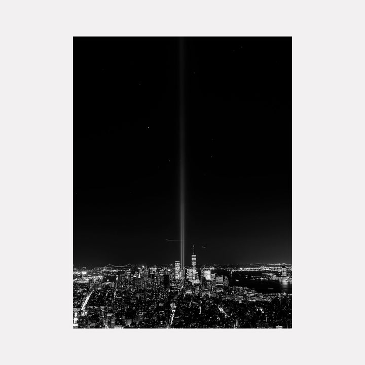 The artwork 'View from Empire State Building' by Garrett Carroll captures a dramatic black and white aerial photograph of Manhattan skyline at night, featuring the iconic Twin Towers memorial lights piercing the dark sky. City lights illuminate the urban grid pattern below, creating a striking contrast between light and shadow across the cityscape.