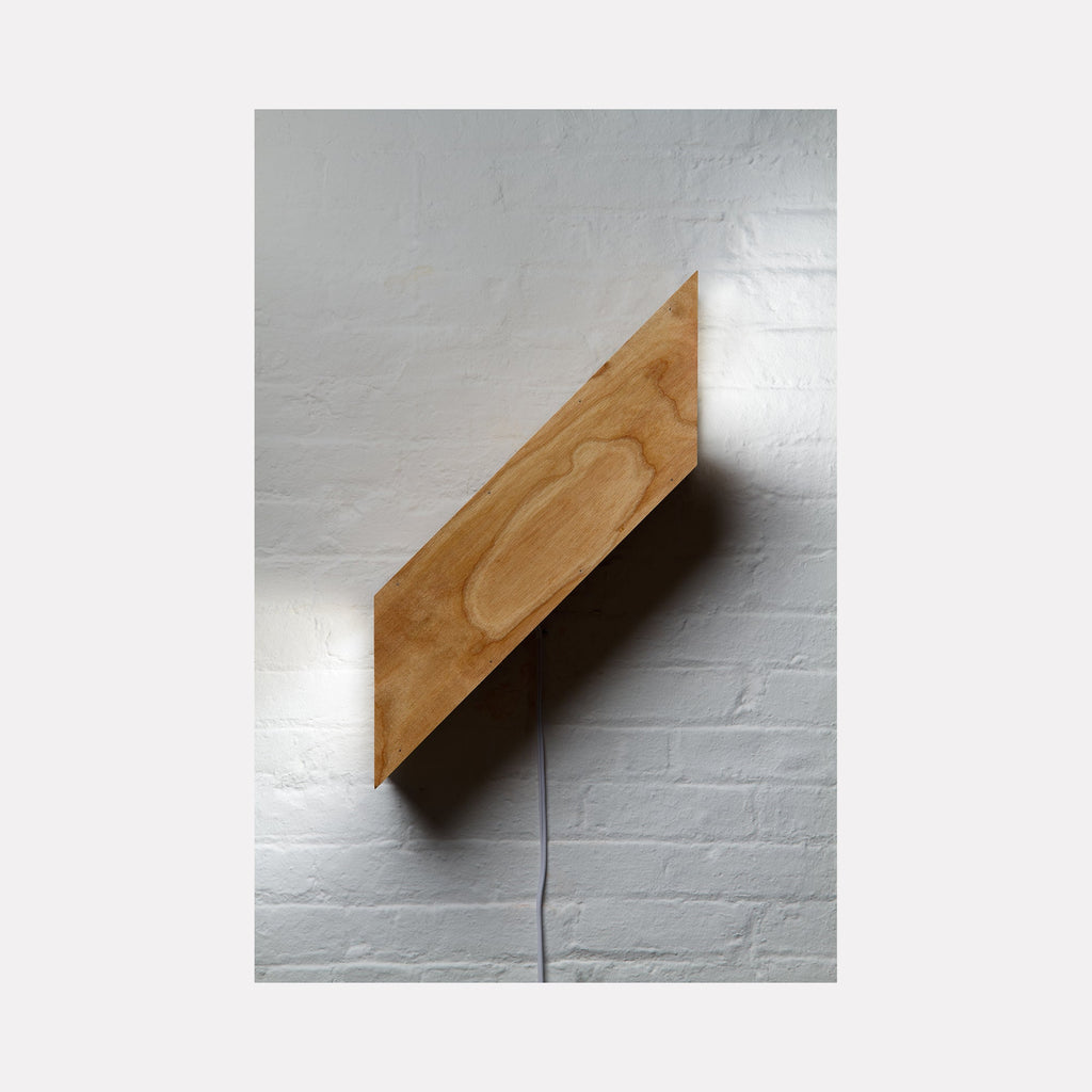 The artwork 'Wood Box (Route 41, Salisbury)' by Ryan Frank features a minimalist light sculpture mounted diagonally on a white brick wall. The wooden box, crafted from plywood, displays natural grain patterns and emits a soft glow through plexiglass, creating a striking contrast against the textured white surface. - Ryan Frank