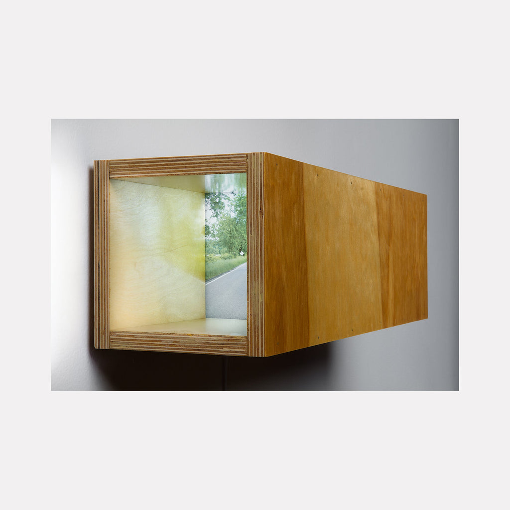 The artwork 'Wood Box (Route 41, Sharon)' by Ryan Frank features a minimalist plywood lightbox mounted on a wall, displaying a luminous photo transparency behind plexiglass. The rectangular wooden structure has layered edges and emits a soft fluorescent glow, creating a window-like view of natural scenery. The honey-toned wood contrasts with the ethereal green-yellow illuminated image. By Ryan Frank.