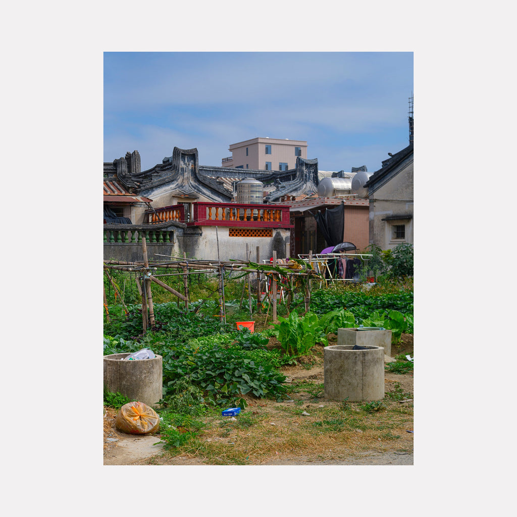 The artwork 'Your Home in Shantou - Series-1' captures a traditional Chinese neighborhood garden with concrete wells, lush green vegetation, and bamboo drying racks against a backdrop of historic buildings featuring distinctive curved rooflines and red temple-style architecture under a soft blue sky. Photography by Garrett Carroll.