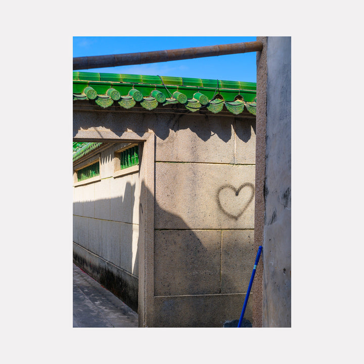 Your Home in Shantou - Series-4 by Garrett Carroll features a traditional Chinese alleyway with emerald green decorative roof tiles, casting shadows on a concrete wall where a heart-shaped shadow appears. The composition captures the contrast between ornate architectural details and simple geometric shadows, with bright blue sky visible above.