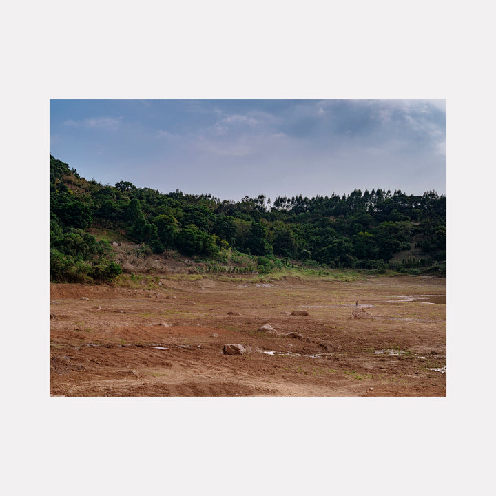 Your Home in Shantou - Series-5 by Garrett Carroll: A contemplative landscape photograph capturing a rustic terrain with reddish-brown earth dotted with scattered rocks, bordered by dense green forest and rolling hills beneath an overcast sky, conveying the raw beauty of undeveloped land in Shantou.