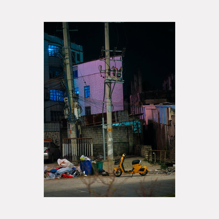 The artwork 'Your Home in Shantou - Series-6' by Garrett Carroll captures a nocturnal urban scene with a pink-colored building, illuminated blue windows, and utility poles dominating the composition. A yellow scooter rests against a concrete wall, while scattered objects and bins line the street, creating a raw, authentic portrayal of city life. The photographic style emphasizes contrasting colors and nighttime ambiance.