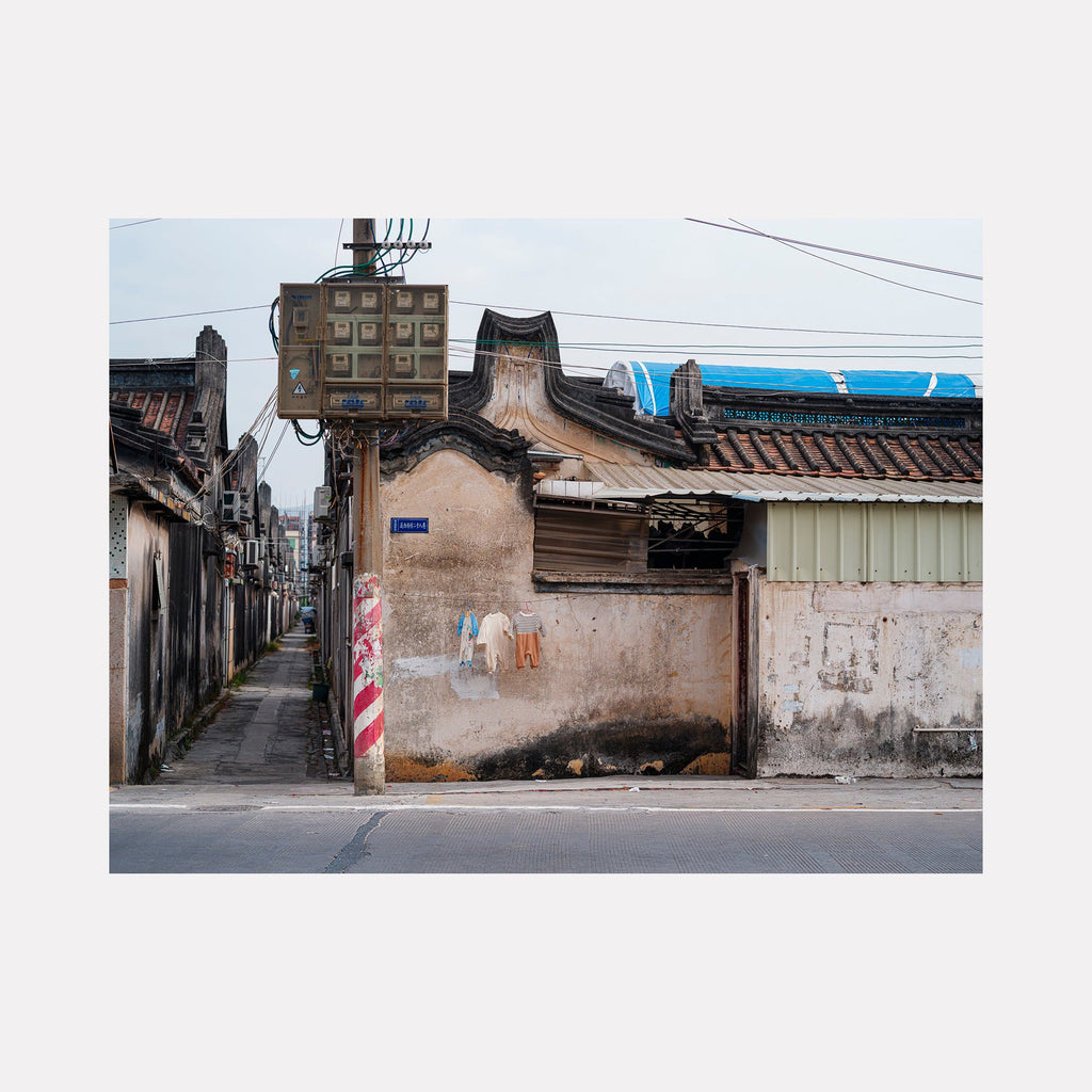 The artwork 'Your Home in Shantou - Series-7' by Garrett Carroll captures a narrow urban alley in traditional Chinese architecture, featuring weathered beige walls, traditional curved rooflines, and vintage electrical boxes. A distinctive red and white striped pole stands against faded concrete, with overhead power lines and blue tarp visible in the composition.