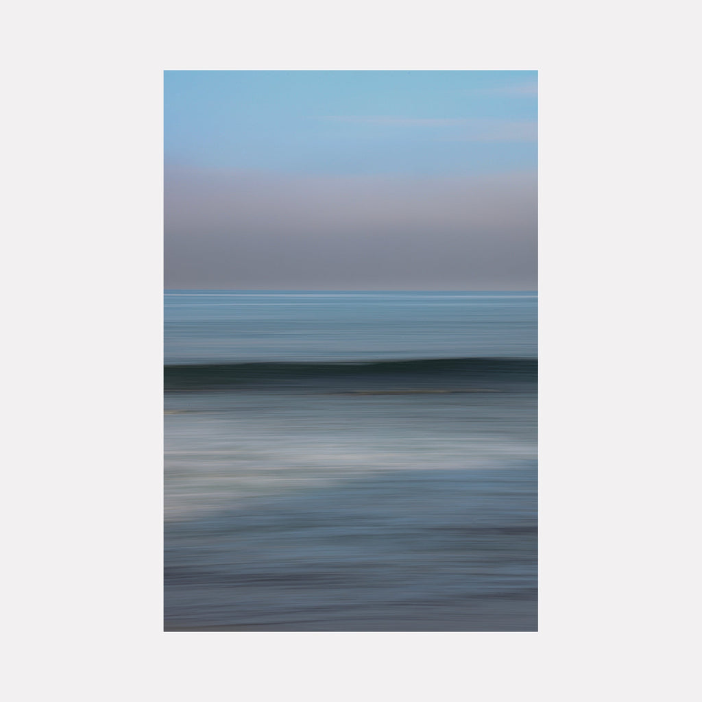 The artwork 'Zuma Wave' by Neil Shapiro captures a minimalist seascape with subtle motion blur, featuring layers of cool blue-grey tones merging into a misty horizon. The abstract composition creates a serene, ethereal effect through horizontal bands of color, showcasing a wave's gentle movement on textured Hahnemuhle print.