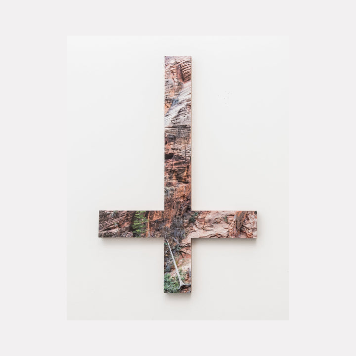 The artwork 'Zion Cross' by Ryan Frank features a striking cross-shaped composition created from a color photograph mounted on sapele wood. The image captures the distinctive red-orange sandstone textures and natural patterns of Zion National Park, with subtle hints of green vegetation integrated into the rugged landscape design.