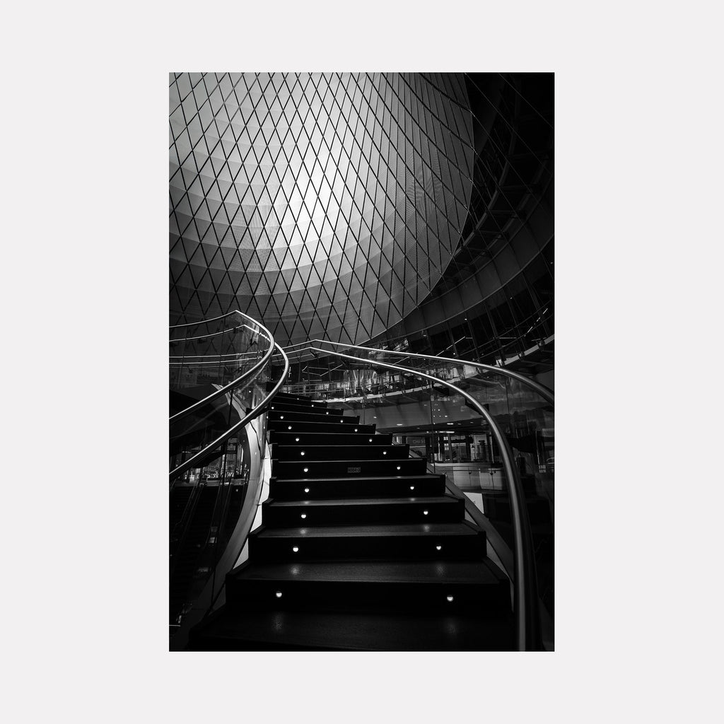 The artwork 'Ascent' by Dennis Maida captures a dramatic black and white architectural photograph of an illuminated spiral staircase, featuring geometric diamond-patterned glass ceiling above. The modernist composition showcases elegant curved handrails, LED-lit steps, and striking contrast between light and shadow, creating a mesmerizing upward perspective.