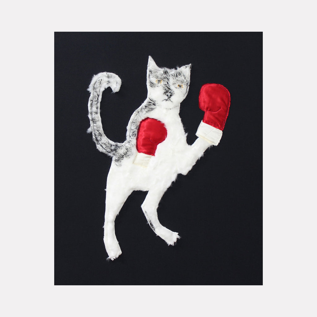 The artwork 'Cat Fight' by Ali Ha features a playful white cat wearing red boxing gloves against a black background, created through collaged fabric applique and acrylic paint. The cat stands upright in a boxing stance, with its tail curved upward, showcasing a whimsical blend of textile art and painted details.